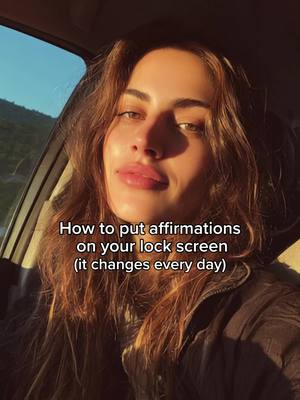 A woman is sitting in a car, looking towards the camera. She has long brown hair. The text overlay reads "How to put affirmations on your lock screen (it changes every day)". The background is lit by the sun. 