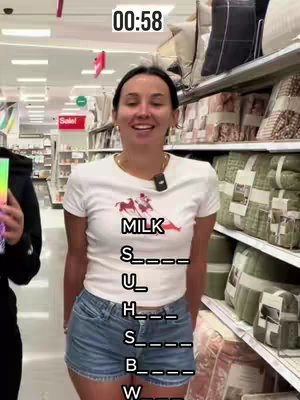 A woman is standing in a store aisle, facing the camera. Text is overlaid on her, partially obscuring her clothing. She is smiling and looking towards the camera. Shelves filled with merchandise are in the background.