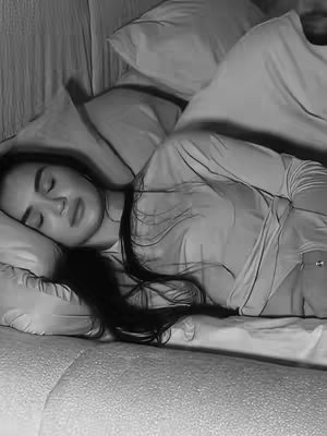 A grayscale image shows a woman lying in bed, seemingly asleep. She rests on a pillow, and pillows are also behind her head. The scene is captured from a slightly high angle.