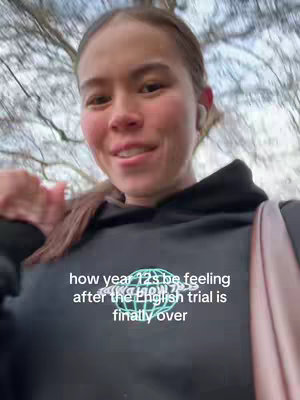 A young woman with brown hair and fair skin is filming herself from a slightly low angle. She is wearing a black hoodie with white text on it, and she has one earbud in. She appears to be outdoors with trees and a cloudy sky visible in the background.