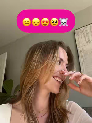 A woman with light brown hair smiles while looking towards the right. She is wearing a white top and has a necklace. Behind her, a picture or art piece hangs on the grey wall. Above her a pink bar contains five emoji faces.