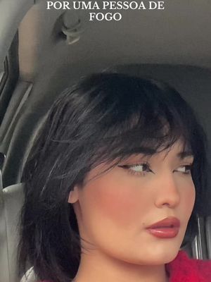 A woman with short, dark hair is seated in a car, with a side-profile view dominating the frame. She wears dark eyeliner and reddish lip color. Her skin is a warm tone with blush applied. The background hints at a gray, slightly blurred interior, with text at the top.