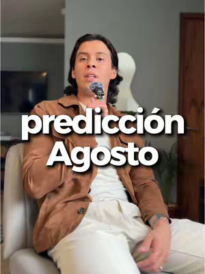 A man with long, dark hair is seated on a light-colored chair. He holds a microphone and appears to be addressing the camera. The overlaid text 'predicción Agosto' is in bold, white font with a black outline. The background includes a gray wall and a glimpse of decor.