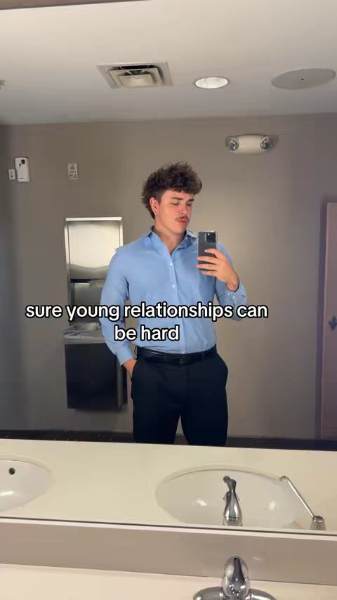 A man is taking a selfie in a public bathroom. He is dressed in a light blue button-down shirt and black pants with a black belt. The person is holding a smartphone and looking at the camera. Behind him is a bathroom with a white sink and a mirror, with a text overplay on the image.