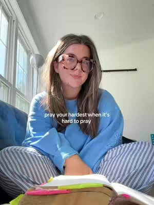 A woman wearing glasses and a blue sweatshirt sits cross-legged on a sofa, holding an open book. The background shows a window and natural light.