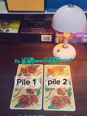 The image features two tarot cards set on a wooden surface, with a small lamp to the right. The text "Pile 1" and "Pile 2" are overlaid on the cards. In the background, there's a collection of books and a shelf.