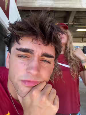 A selfie-style image featuring a young man in the foreground with his arm resting on his chin, slightly gazing downwards. He is wearing a maroon t-shirt and a silver chain. Behind him, partially visible, is a woman with blonde hair, wearing a red hat and a red shirt, and is slightly out of focus. The background suggests an outdoor stadium setting with concrete structures. The overall lighting seems natural.