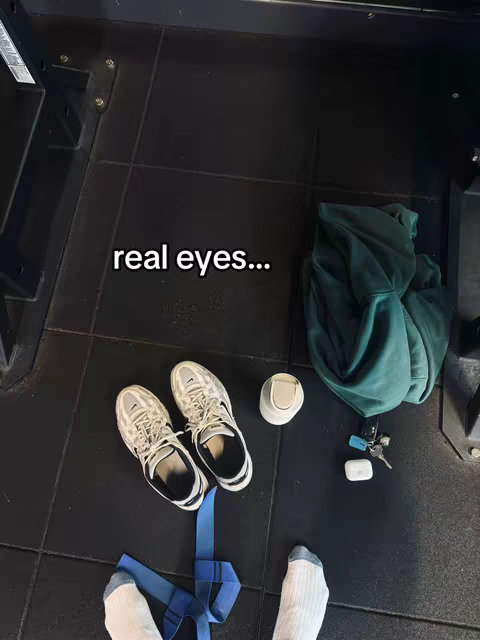 An overhead shot shows a gym floor with various items scattered around. There are a pair of athletic shoes, a green hoodie, keys, a bottle, a blue exercise band, and a pair of white socks. The floor is black.