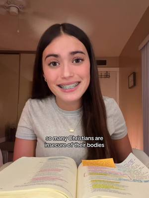 A young woman with long brown hair and braces is smiling at the camera. She is wearing a grey t-shirt and a necklace. She's sitting behind an open book. Text overlay reads, "so many Christians are insecure of their bodies". The background includes a ceiling fan, a partially visible room and a window.
