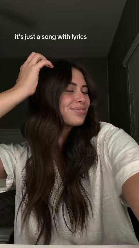 A woman with long dark hair is shown from the chest up against a shaded backdrop. She wears a white top and appears to be singing with her eyes closed. Text overlayed at the top reads "it's just a song with lyrics".