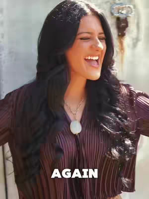 A woman with long black hair, wearing a purple top, smiles widely with her head tilted slightly back. The background is blurred but seems to be a light-colored surface. Text at the bottom reads "AGAIN".