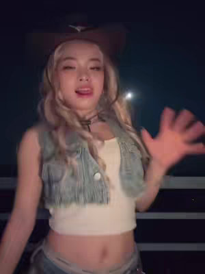 A woman with long, wavy blonde hair and a cowboy hat is smiling and gesturing with her right hand. She wears a denim vest over a white top, and denim pants or shorts, with a dark night background.