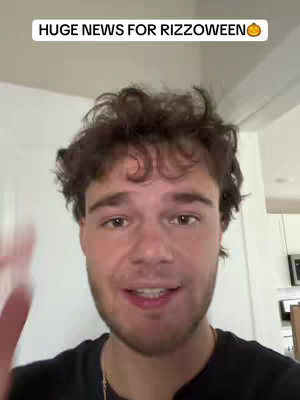 A young man with short, curly brown hair looks toward the viewer and speaks in a room. A text overlay at the top states "HUGE NEWS FOR RIZZOWEEN", with a pumpkin emoji. The man wears a black t-shirt and a gold necklace and gestures with his right hand. Behind him, a white wall, a door frame, and a glimpse of a dark object are visible. The lighting seems natural.