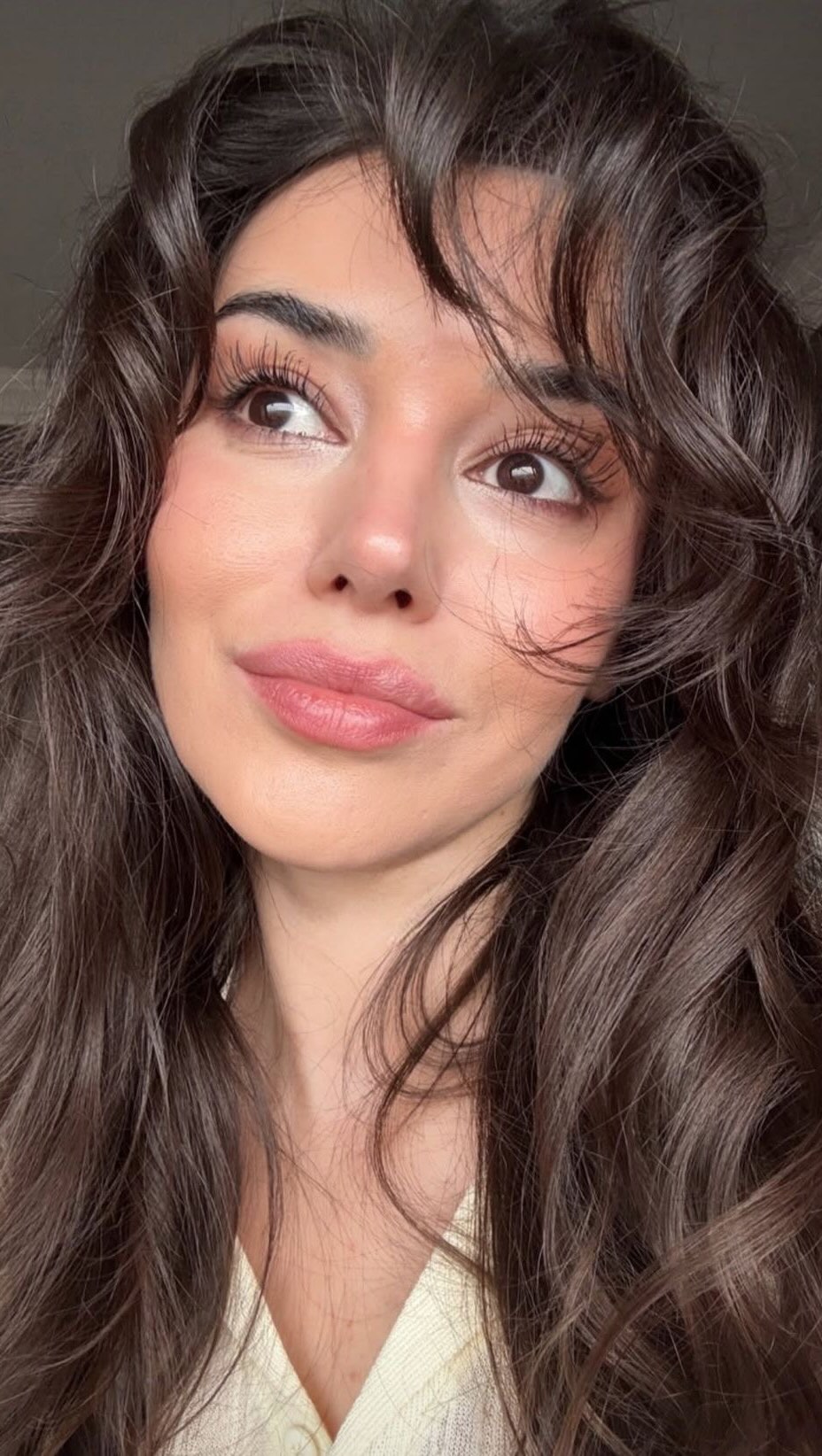 A close-up shot of a woman's face with long, wavy brown hair. She is looking slightly upwards and to the right, with a soft smile on her face. Her skin appears smooth and has a natural glow, with visible makeup including blush, lipstick and mascara. The lighting is soft, creating a flattering effect.