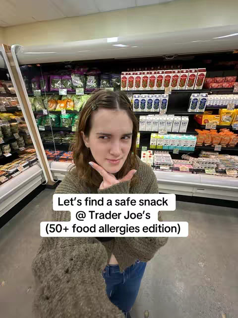 A woman is standing in a brightly lit grocery store aisle. Cool-toned fluorescent lights illuminate the shelves filled with a variety of packaged goods. The woman, with light skin and dark hair, faces the camera and holds up her fingers. Text overlays create a frame with information about the video’s topic.