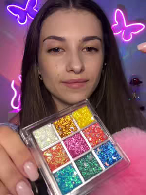 A young woman with long brown hair and a neutral facial expression holds up a clear, square palette of nine colorful glitter eyeshadows. Behind her, the background features purple and pink neon butterfly lights. She holds a fuzzy pink item in her other hand, and her nails are painted pink.