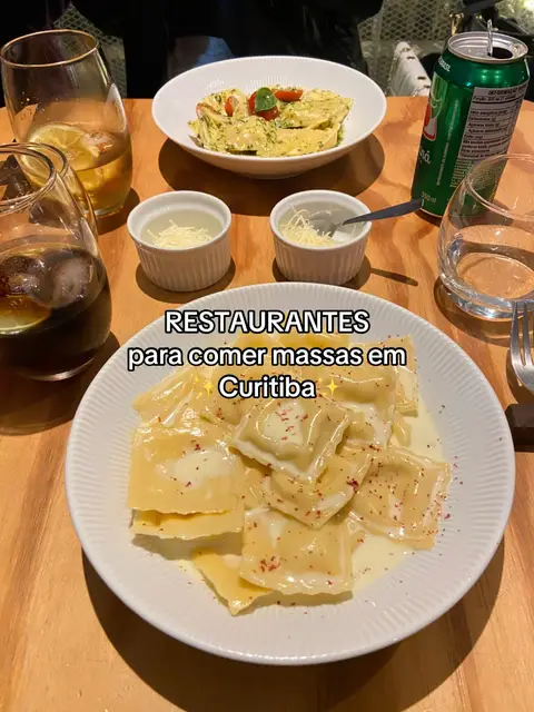A top-down shot of a wooden table set for a meal. On the table are two plates of pasta dishes, small bowls of grated cheese, glasses with drinks, and a can of soda. Text overlay at the bottom says, "RESTAURANTES para comer massas em Curitiba"