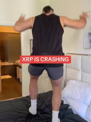 A man standing on black sheets on a bed, jumping up in the air with his arms spread. A red rectangular bar with the text "XRP IS CRASHING" is in the center of the image. The background shows a room with a door, a window, and a bathroom area.