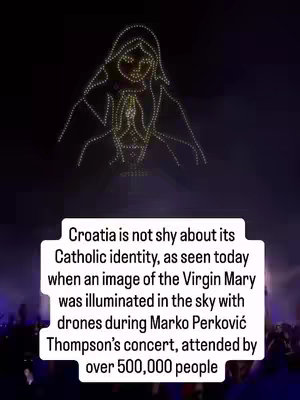 The image shows a night sky display featuring a light-based rendering of the Virgin Mary above a crowd. The Virgin Mary is depicted in the form of a figure constructed from illuminated points or lights, with hands clasped in prayer. Below, the bottom of the image shows a crowd of people, suggesting a public event or concert. The image has a dark background with the illuminated figure as the focal point. A rectangular text overlay is at the bottom of the image with details about the event.