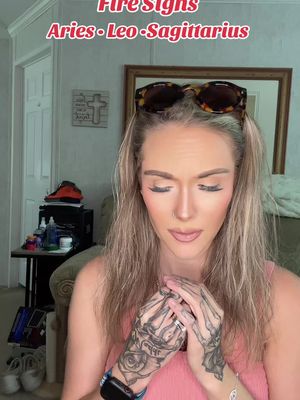 A woman with long blonde hair and sunglasses on her head looks down with a slightly sad expression. She has visible hand tattoos. The image has text overlaying it that states 'Fire Signs Aries Leo Sagittarius'.