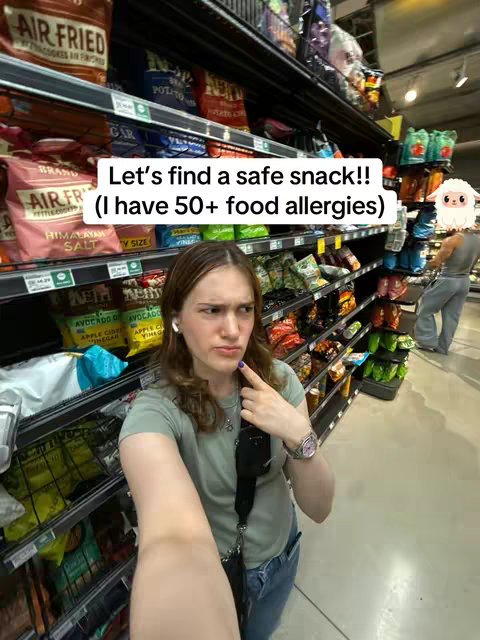 A woman is inside a grocery store, taking a selfie. She has a concerned expression and points at her chin with a finger. The background consists of packed shelves of snacks and other food items. Text overlays in the image provide context about food allergies.