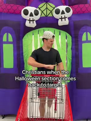A man in a baseball cap and black shirt is inside of a red shopping cart in front of a Halloween-themed inflatable display, pointing to the side with his right hand. Above him are two skull decorations on top of purple structure and green cutout.