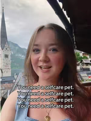 A woman with long blonde hair is talking to the camera, framed against a backdrop of a European town with a church steeple, mountains, and a cloudy sky.
