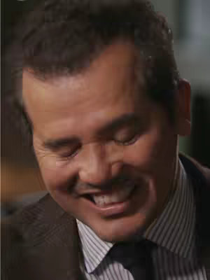 A close-up shot of a smiling man with dark hair and a mustache, wearing a suit and tie. His eyes are closed, and he has a warm, genuine expression.