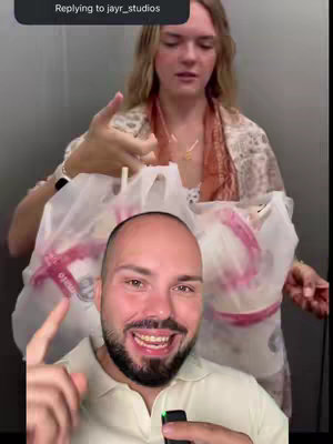 A selfie-style video cover featuring a man in the foreground, smiling at the camera and pointing a finger towards himself, while a woman holding what appears to be bags of food is visible in the background. The setting looks like an elevator.