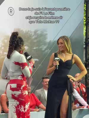 The image shows two women with microphones, seemingly at an event. The woman on the left has a patterned red and white dress, and her hair is styled in a braid. The woman on the right is wearing a black dress with a high slit, her hair is styled in waves. They are both smiling and looking at each other.