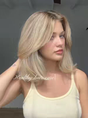 A woman with long blonde hair is looking at the camera. She is wearing a light yellow tank top. The background is blurry and appears to be a wall.