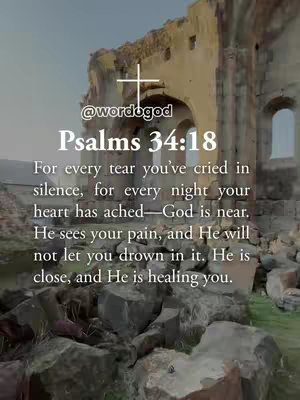 The image is a vertical shot depicting a weathered building with an open archway. A white cross is overlaid, along with the handle '@wordogod' and the text of a Bible verse, Psalms 34:18, in a sans-serif font. The verse begins with 'For every tear...'. The color palette consists of muted earthy tones derived from the stone structure and the cloudy sky, providing a backdrop of calm and reflection. The layout is straightforward, with centered text that guides the eye. Rusted, rough stones fill the foreground.