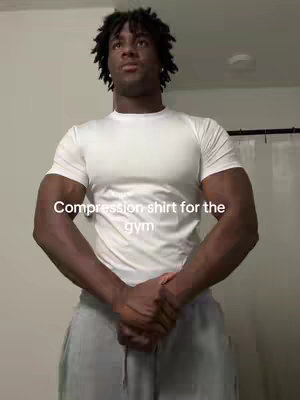 A man with dark skin and short, curly hair is centered in the frame from the chest up in a well-lit room. He wears a white compression shirt and gray sweatpants. The image is framed vertically, with the man's arms crossed over his chest, hands clasped. The immediate background is a wall and door frame. Text overlayed on the image reads, 'Compression shirt for the gym'.