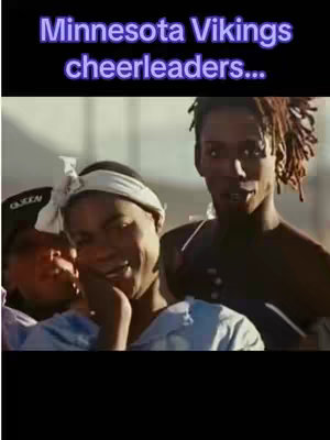 The image is a still from a video, featuring three individuals with dark skin. The central figure, in a light-colored headband and a light blue top, is smiling with his hand near his face. To his right, stands another individual with styled hair, slightly out of focus. The top of a black cap with the word "QUEEN" is partially visible on the left. The text overlay at the top reads "Minnesota Vikings cheerleaders..." in a purple font.