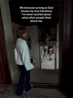 A woman stands inside a building, facing away from the camera. A text overlay is positioned above her head, and she's looking towards a mirror or reflective surface, which shows a glimpse of a hallway with blurred figures.
