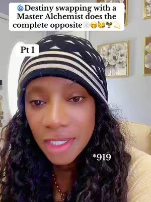 A selfie-style video cover image featuring a Black woman with long, wavy dark hair, wearing a black and white patterned beanie. She's looking directly at the camera with a slightly open mouth, as if speaking. The background is a blurry interior scene with framed artwork visible. Text overlays include the title 'Destiny swapping with a Master Alchemist does the complete opposite' at the top, and 'Pt 1' at the lower left, with the number '*919' closer to the bottom right.