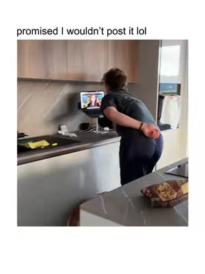 A man stands in a kitchen, facing a laptop on the counter. The laptop screen displays two people, likely for a video call. He is wearing a dark shirt and pants. The background is a modern kitchen with wooden cabinets and a countertop. A bag of snacks sits on the counter.