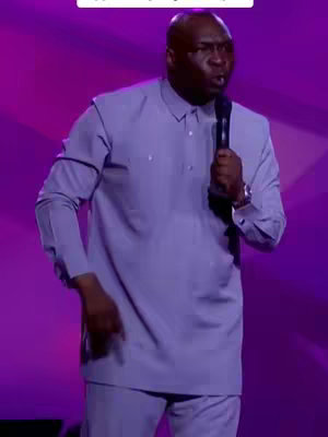 A man in a light blue shirt and pants stands on a stage, holding a microphone. The background is a gradient of pink and purple.