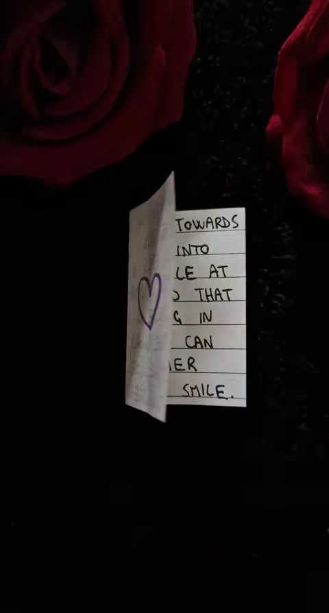 The image shows a close-up of a handwritten note with a heart drawn on the folded section. The note is partially folded, revealing a heart drawn with purple ink and handwritten text that reads "TOWARDS INTO LE AT J THAT G IN CAN ER SMILE." The background has two red roses and a black textured surface.