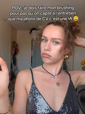 A young woman with blue eyes and light skin is taking a selfie while looking at the camera with a somewhat puzzled or unhappy expression. The image is framed from the chest up, with the woman's short curly hair and the top of a tank top visible in the shot. Text overlayed on the image reads in french.