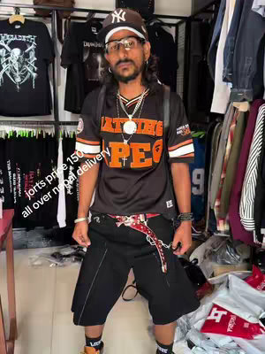A man stands in front of racks of clothing, wearing a black jersey with orange lettering, black shorts, and a baseball cap. The image has text overlays advertising shorts for price 150 and all-over Nepal delivery. The background shows clothing racks filled with various items.