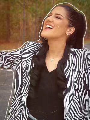 A woman with dark hair and dark eyes stands outdoors with a blurred background, the woman is angled up and to the left with her mouth open, revealing teeth in an expression of joy. She has a zebra-striped jacket over a black top. She has a cross necklace.