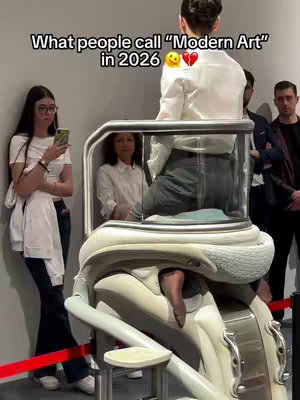 A woman in a white dress is seated inside a glass-enclosed, futuristic-looking structure that resembles a medical device. A young woman in a white top and black pants with glasses is observing and holding a phone. The image is taken in an art gallery, and there are other people are in the background.