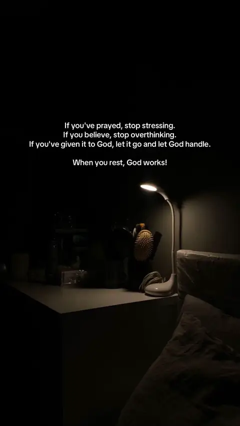 The image is a dimly lit interior scene, likely a bedroom, with a bedside table and a bed partially visible. A desk lamp is the main light source, illuminating the table and a portion of the wall. On the table, there is an array of objects, including what appears to be a decorative brush and other items obscured by shadows. Text is overlaid on the image, formatted in white and centered, with the core message focusing on trust and letting go. The overall composition is minimalistic, with a dark aesthetic that creates a sense of contemplation and serenity.