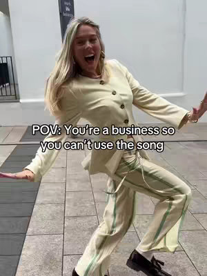 A woman in a cream-colored suit is captured in a joyful pose outdoors, her arms outstretched and mouth open in an expression of delight. The background is a lightly blurred urban setting with a grey tiled pavement and glimpses of building exteriors. Text overlaid on the image reads, 'POV: You're a business so you can't use the song'.