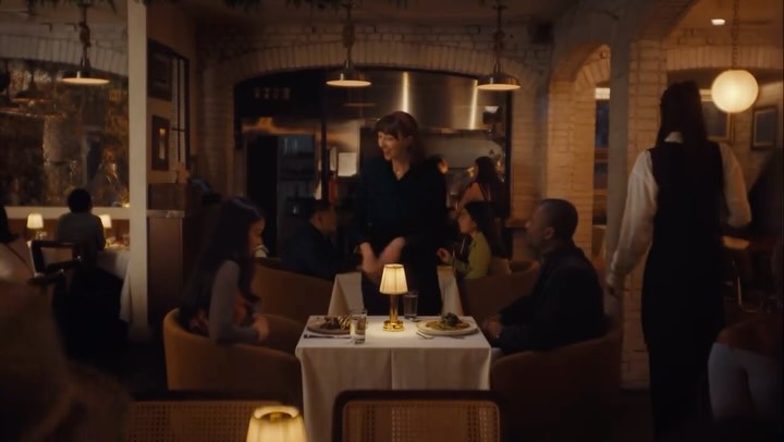 A well-lit restaurant interior. A waitress is approaching a table with served meal and a small table lamp. People are sitting at other tables.