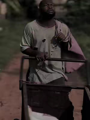 The image shows a man with a dark complexion, dressed in a worn, light-colored t-shirt and pants, pushing a metal cart. He is looking up and to the side with a contemplative expression. The background is slightly blurred, suggesting an outdoor setting, perhaps a rural or underdeveloped area. The overall tone seems somber, possibly depicting a scene of hardship or introspection.