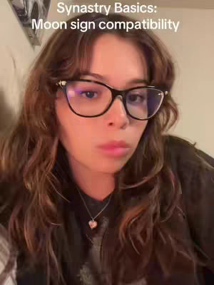 A woman with long brown wavy hair is centered in the frame. She wears black-rimmed glasses and a black top. Text at the top reads: 'Synastry Basics: Moon sign compatibility'.