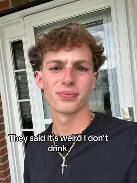 A young man with short, curly brown hair stands in front of a white door and window frame, looking at the camera with a neutral expression. He's wearing a black t-shirt and a gold chain with a cross pendant. Text overlaid on the image reads, "They said it's weird I don't drink.