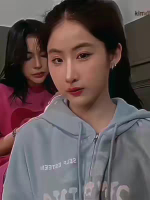 Two young women are shown in a selfie-style shot. The woman on the right is the main focus; she wears a light blue hoodie, looks directly at the camera, and has a slightly serious expression. The woman on the left appears slightly behind and to the left, wearing a pink shirt and focused on the person taking the photo. The overall lighting is natural, and the background is out of focus, giving a sense of intimacy.